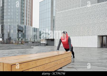 Young sporty Caucasian woman doing push-ups on wooden bench while having outdoor workout in downtown on cold autumn morning Stock Photo