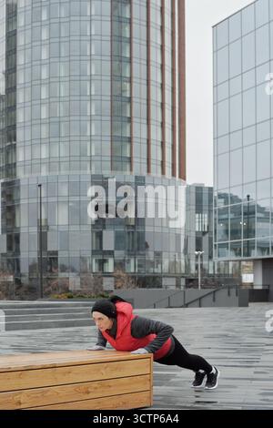 Young athletic Caucasian woman in warm activewear doing push-ups on wooden bench while having workout in downtown on gloomy afternoon Stock Photo