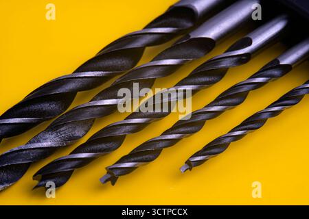 Assorted drill bits on a yellow background. Close-up showcases a set of various-sized metal drill bits arranged diagonally on a bright yellow surface. Stock Photo