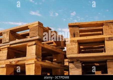 Stacked wooden shipping pallets under blue sky. Low-angle close-up of rough-sawn wooden pallets stacked outdoors. Suitable for themes like logistics. Stock Photo