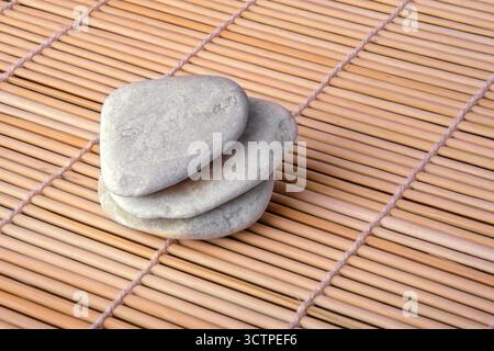 A small stack of three smooth, flat grey pebbles arranged neatly on a bamboo mat. The contrast between the cool stones and the warm wooden texture. Stock Photo