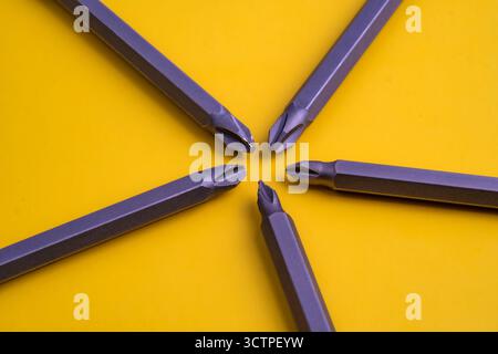 Close-up of five metallic screwdriver bits arranged in a star-like formation on a yellow background. The sharp cross tips highlight precision. Stock Photo