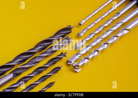 Collection of various metal drill bits, both dark and shiny are arranged diagonally on a vibrant yellow background. Showcasing different sizes. Stock Photo