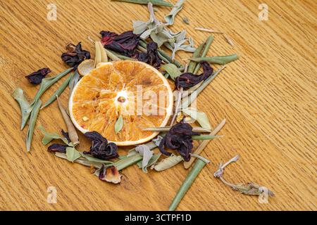 Dried a orange slice is centrally placed on wooden surface. Surrounded by an assortment of dried botanicals including dark red hibiscus flowers. Stock Photo