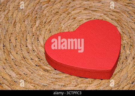 Red heart-shaped paper box placed on a woven straw mat. Symbol of love, care and affection with rustic background and simple romantic decoration. Stock Photo