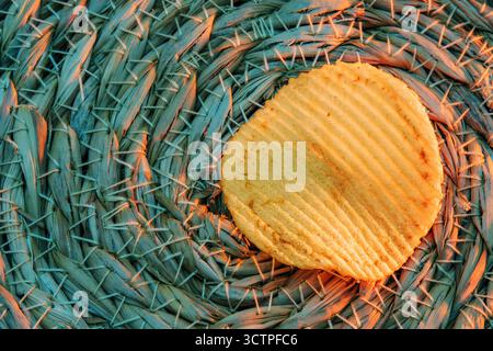 Potato chip on textured background . Single ridged potato chip, illuminated by golden light, rests on a textured. Woven mat of natural fibers. Stock Photo