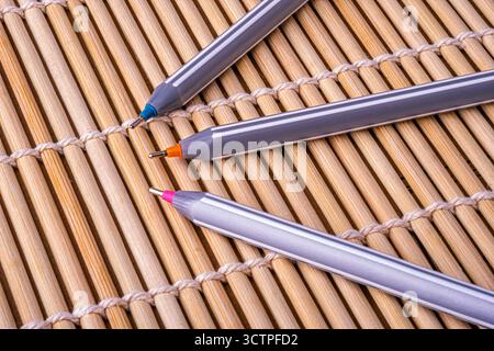 Three ballpoint pens with colorful tips  are placed diagonally on bamboo mat. The natural wooden texture contrasts with the modern stationery design. Stock Photo