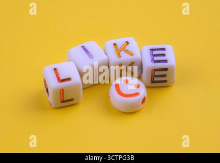 White cubes with colorful engraved letters spelling "LIKE" placed on a bright yellow background. Accompanied by a bead with an orange smiley face. Stock Photo