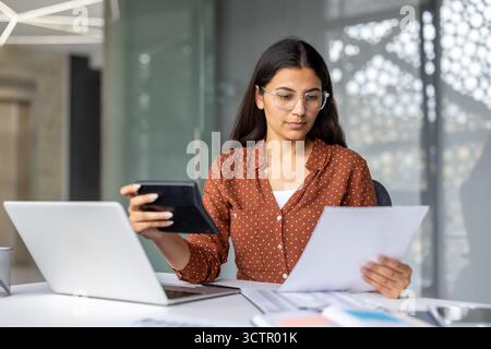 Indian businesswoman in modern office reviewing financial reports, using laptop and calculator while focused on budgets, accounting and investment documents at her desk Stock Photo