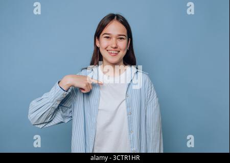 Young woman smiling and pointing at herself against blue background. Stock Photo