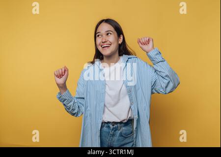 Happy young woman in casual outfit dancing against yellow background. Stock Photo