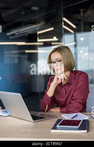 Businesswoman wearing glasses concentrating on laptop screen, analyzing data and thinking about solutions while sitting at her desk in a modern corporate office Stock Photo