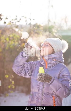 Girl in warm clothes playing with soap bubbles during cold sunny winter day, representing innocence and simple happiness. Stock Photo