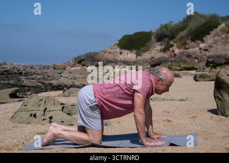 Elderly man practicing yoga on the beach doing cat pose for back health Stock Photo