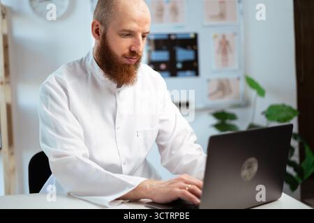 Bearded Male Doctor wearing white coat in Hospital Office using Laptop computer, Analyzing Medical Data. Stock Photo