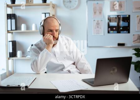 Tired lazy Bearded Male Doctor wearing white coat in Hospital Office using Laptop computer, Analyzing Medical Data. Stock Photo