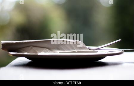 Close-up view of a clean, minimalist table setting with a folded white napkin and a stainless-steel fork resting on a white plate. Stock Photo