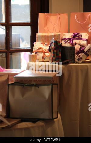Collection of wrapped gift boxes and colorful gift bags arranged on a table near a window, illuminated by warm natural light at a celebration or event Stock Photo