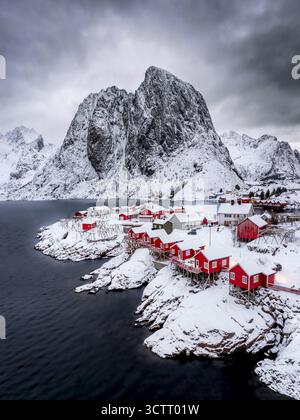 Aerial view of red cabins nestled on the snow-dusted shores beneath the towering, rugged peaks of Hamnoy, a stark contrast against the dark, rippling waters, Hamnoy, Nordland, Norway. Stock Photo