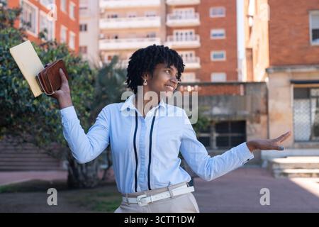Happy young woman smiling, holding a book and wallet in a city square Stock Photo