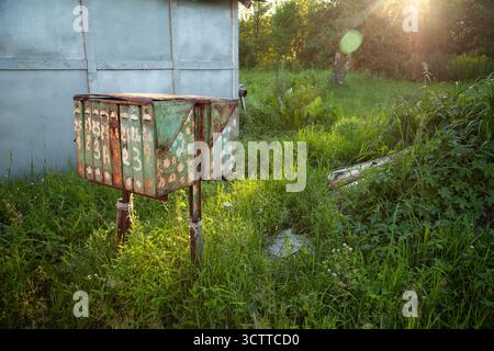 An old, rusty postbox stands weathered in a quiet Russian village, surrounded by green grass. A group of worn metal mailboxes with peeling paint Stock Photo