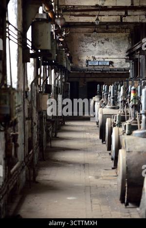 An abandoned corridor in an old, decaying factory hall with a long row of dusty, decommissioned machines Stock Photo