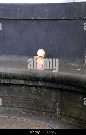 An open, half-eaten can of Maggi ravioli as an urban still life, left behind on a wet, dark stone bench in a city. Stock Photo