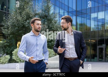 Two male businessmen actively engaging in a focused conversation, discussing important work matters and business development outside a contemporary glass office building Stock Photo