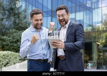 Two happy businessmen celebrating an achievement by clenching their fists and shouting with joy while ,looking at a digital tablet screen outdoors in front of a modern office building Stock Photo