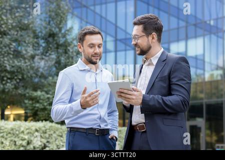 Two businessmen collaborate outdoors, reviewing ideas and data on a tablet in front of a modern glass office building, discussing strategy, planning and partnership Stock Photo