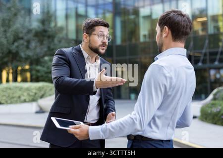Two angry businessmen standing outdoors, having a heated argument about business issues, with one man using a digital tablet during the intense professional discussion Stock Photo