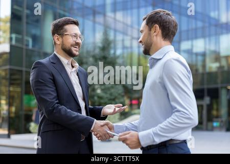 Two smiling professional businessmen are shaking hands, confirming a successful business deal or partnership in an urban setting with a modern office building in the background Stock Photo