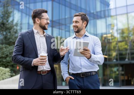 Two cheerful businessmen stand by a modern office building, holding a tablet and coffee, smiling and chatting outdoors during a casual work break about strategy and collaboration Stock Photo