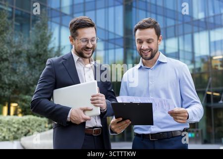 Two smiling adult businessmen, one holding a laptop and the other reviewing documents on a clipboard, are standing and discussing business matters outdoors in front of a modern corporate building Stock Photo