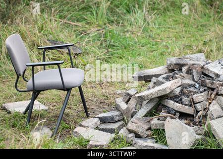 Old chair near extinguished stone fire pit on green lawn, empty abandoned outdoor place without people Stock Photo