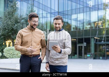 Two professional businessmen discussing a new project, collaborating and sharing ideas while looking at a digital tablet screen outside a modern office building Stock Photo