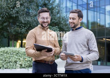 Two smiling male colleagues discussing business and sharing ideas while reviewing information on a digital tablet and clipboard outside a modern building Stock Photo