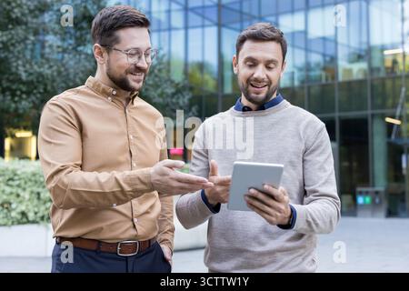 Two smiling adult businessmen engaged in a dynamic discussion, sharing ideas and collaborating on a project using a digital tablet outside a modern office building Stock Photo