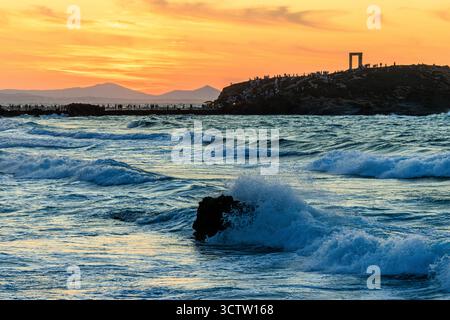 waves breaking on rocks in the bay in front of tourist crowds leaving portara giant doorway of Naxos framed against yellow sunset sky Stock Photo
