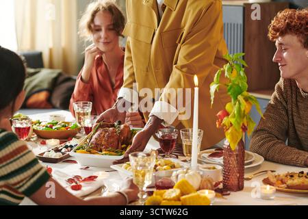 Young Black Man Serving Roast Turkey to Diverse Young Adults at Dinner Table Stock Photo