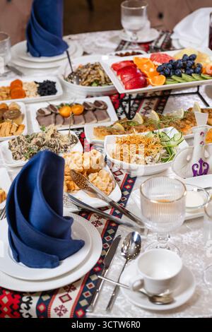 A richly set table featuring assorted appetizers, fresh fruit platter, crispy pastries, and elegantly folded navy blue napkins on crisp white plates. Stock Photo