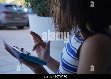 A close-up, candid shot of a girl playing a Nintendo Switch handheld game console outdoors while holding a small note, featuring a blue and white top. Stock Photo