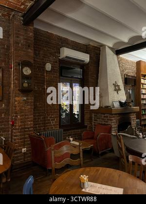 The photo shows an indoor seating area with brick walls, a white plaster fireplace, red armchairs, wooden chairs, and round tables. A window with nigh Stock Photo