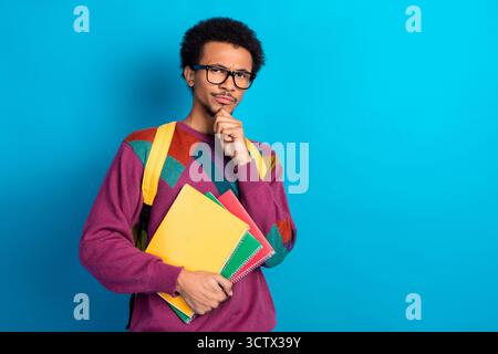 Confident young man holding colorful notebooks against a vibrant blue background, wearing a trendy sweater Stock Photo