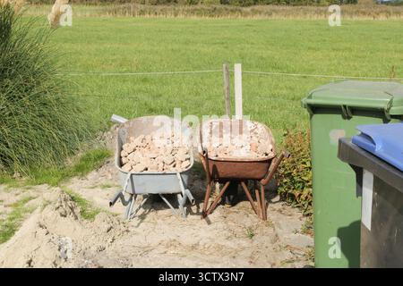 two wheelbarrows with rocky debris and a waste bin are standing outdoor during works at a construction site for a new shed in the countryyside Stock Photo