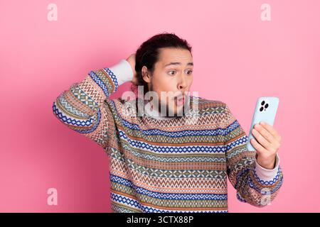Surprised man wearing colorful sweater looking at smartphone on pink background, expressing shock and excitement Stock Photo