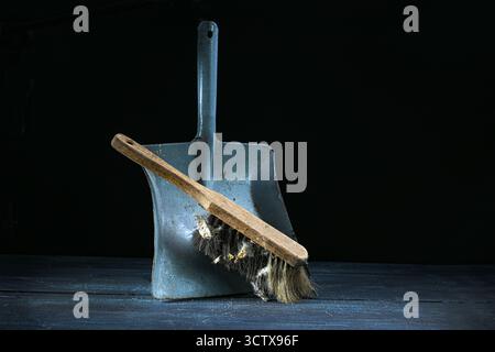 Old hand brush with wooden handle and a lot of dirt leaning on an aged metal dustpan against a dark background, still life of vintage cleaning tools, Stock Photo