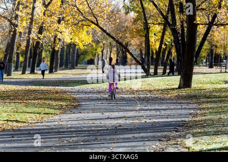 Child riding a pink bicycle along a tree-lined path in Bowness Park during autumn with colorful foliage and people walking. Calgary Alberta Canada Stock Photo