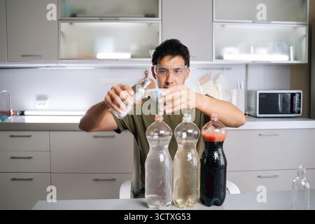 Portrait of focused chemist wearing safety glasses carefully pouring liquid from glass bottle through funnel into plastic bottle, creating plant Stock Photo