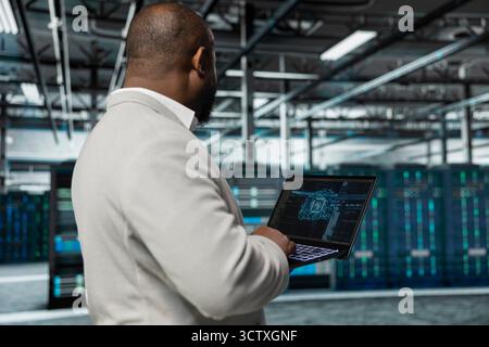 Man typing on laptop in server hub, monitoring data center energy consumption using AI software. Engineer managing supercomputers in server farm using artificial intelligence tech Stock Photo